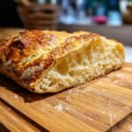 Close-up of a freshly baked loaf of artisan bread, sliced to reveal its airy crumb and golden crust, on a wooden cutting board.