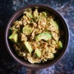 A close-up overhead view of Creamy Avocado Tuna Salad with chunks of avocado and sprinkled red pepper flakes in a dark bowl.