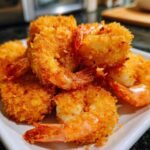 A close-up of golden-brown, crispy panko shrimp piled on a white plate, showcasing their crunchy coating and visible tails.