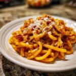 Close-up of a plate filled with bucatini pasta in a rich tomato sauce, topped with grated cheese and black pepper.