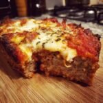 A slice of Delicious Traeger Pizza Meatloaf Recipe For Families, topped with melted cheese and marinara sauce on a wooden board.