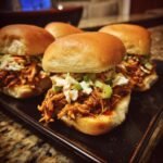 Close-up of Flavorful BBQ Chicken Sliders topped with coleslaw and green onions on a dark tray.