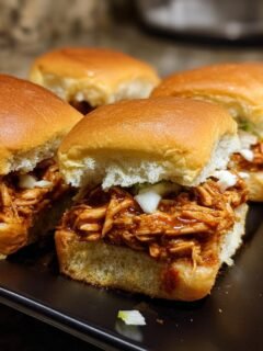 Close-up of four Flavorful BBQ Chicken Sliders on a black plate, topped with diced onions and coleslaw.