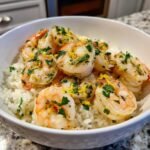 A close-up of a white bowl filled with fluffy white rice topped with succulent lemon garlic shrimp and fresh parsley.