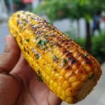 A hand holding a perfectly grilled corn on the cob, seasoned with herbs and spices.