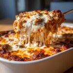 A serving spoon lifts a portion of Irresistible Baked Ziti With Sausage, showcasing gooey melted cheese strings.