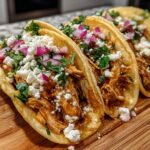 Close-up of three Irresistible Chicken Tacos With Creamy Chipotle Sauce, topped with crumbled cheese, red onion, and cilantro.