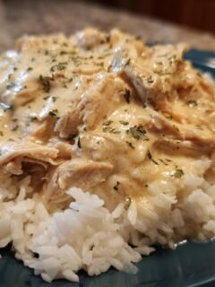 A close-up of Irresistible Crockpot Crack Chicken and Rice on a blue plate, featuring shredded chicken in a creamy sauce over white rice, garnished with parsley.