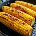 Close-up of four ears of Irresistible Grilled Corn On The Cob, perfectly charred and glistening in a grill pan.