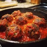 Close-up of Irresistible Smoked Italian Meatballs simmering in a rich tomato sauce in a cast-iron skillet.