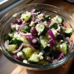 A close-up of a refreshing cucumber feta salad in a glass bowl, featuring chopped cucumbers, feta cheese, red onion, and olives.