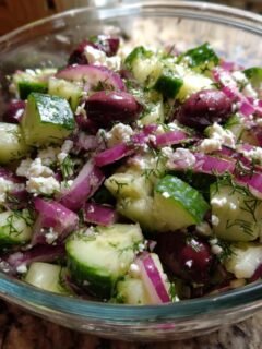 A close-up of a refreshing cucumber feta salad in a glass bowl, featuring chopped cucumbers, red onion, feta cheese, olives, and dill.