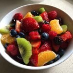 A close-up of a white bowl filled with a Vibrant Fresh Fruit Salad, featuring strawberries, blueberries, raspberries, kiwi, and orange slices.