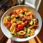 A bowl of zesty pasta salad with Italian dressing, featuring shell pasta, cherry tomatoes, cucumber, olives, and red peppers.