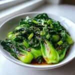 A close-up of vibrant green, stir-fried Baby Bok Choy coated in a dark savory sauce and sprinkled with sesame seeds.