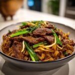 Close-up of a hearty bowl of Beef Chow Fun topped with sliced beef and bright green scallions.