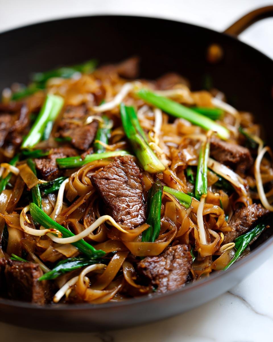 Close-up shot of savory Beef Chow Fun noodles, beef slices, and green onions in a dark wok.