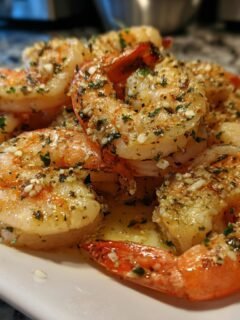 Close-up of juicy Baked Shrimp Scampi piled on a white plate, glistening with garlic butter and herbs, with a lemon wedge.