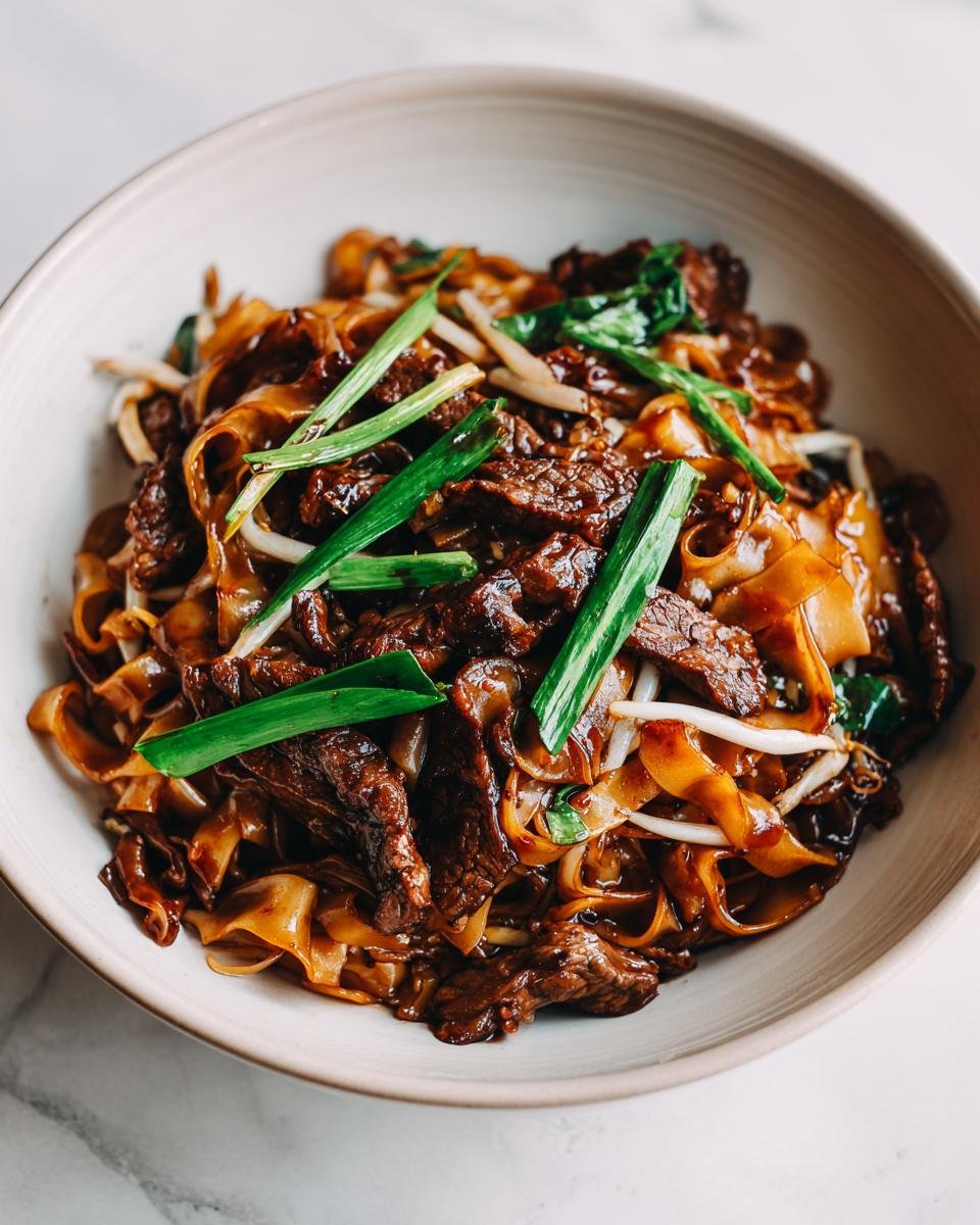 A close-up of perfectly cooked Beef Chow Fun featuring wide noodles, dark sauce, tender beef strips, and green onion garnish.