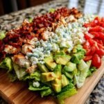 A vibrant, deconstructed Cobb Salad featuring rows of lettuce, grilled chicken, bacon, blue cheese, avocado, and tomatoes.