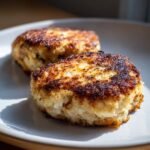 Two perfectly browned and crispy Air Fryer Fish Cakes resting on a light-colored plate, illuminated by sunlight.