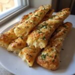 A stack of golden, cheesy Easy Garlic Parmesan Breadsticks sprinkled with fresh parsley on a white plate.