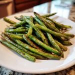 A close-up of glossy, slightly charred Roasted Green Beans piled on a white plate.
