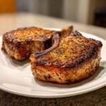 Two thick, bone-in Ranch Pork Chops with a beautiful dark sear and seasoning resting on a white plate.