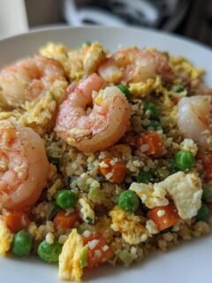 Close-up of a white plate piled high with Shrimp Fried Cauliflower Rice, featuring large pink shrimp, scrambled egg, peas, and carrots.