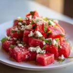Close-up of vibrant, cubed watermelon salad topped with crumbled feta cheese and fresh mint leaves.