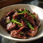 Close-up of dark, glossy Mongolian Beef strips coated in sauce and topped with bright green sliced scallions.