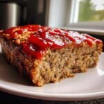 Close-up of a thick slice of juicy Air Fryer Meatloaf topped with a bright red ketchup glaze on a white plate.