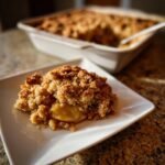 A close-up serving of warm Apple Crumble with a thick, golden, crunchy topping on a white square plate.