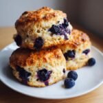 Three golden-brown Blueberry Scones stacked and arranged on a white plate with fresh blueberries scattered nearby.