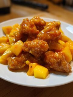 Close-up of glazed, crispy Mango Chicken pieces mixed with fresh, diced mango chunks on a white plate.