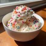 Three scoops of creamy white Birthday Cake Ice Cream generously topped with colorful rainbow sprinkles in a white bowl.
