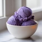 Three scoops of rich, purple Blueberry Nice Cream served in a white bowl on a marble surface.
