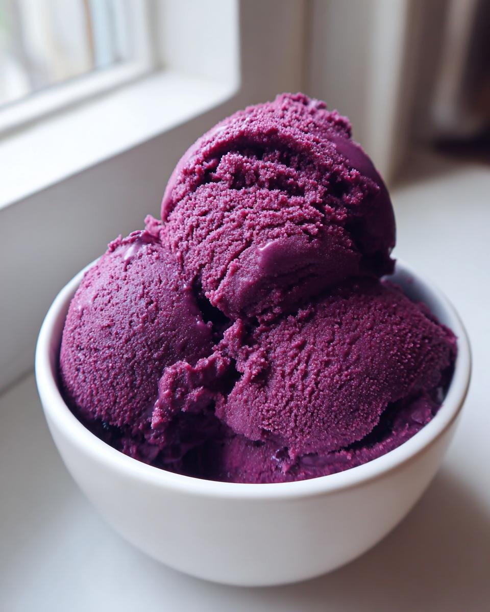 Three scoops of vibrant, deep purple Blueberry Nice Cream served in a white bowl near a window.