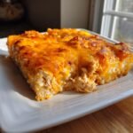 A close-up of a square serving of Buffalo Chicken Rice Casserole topped with bubbly, melted orange cheddar cheese.