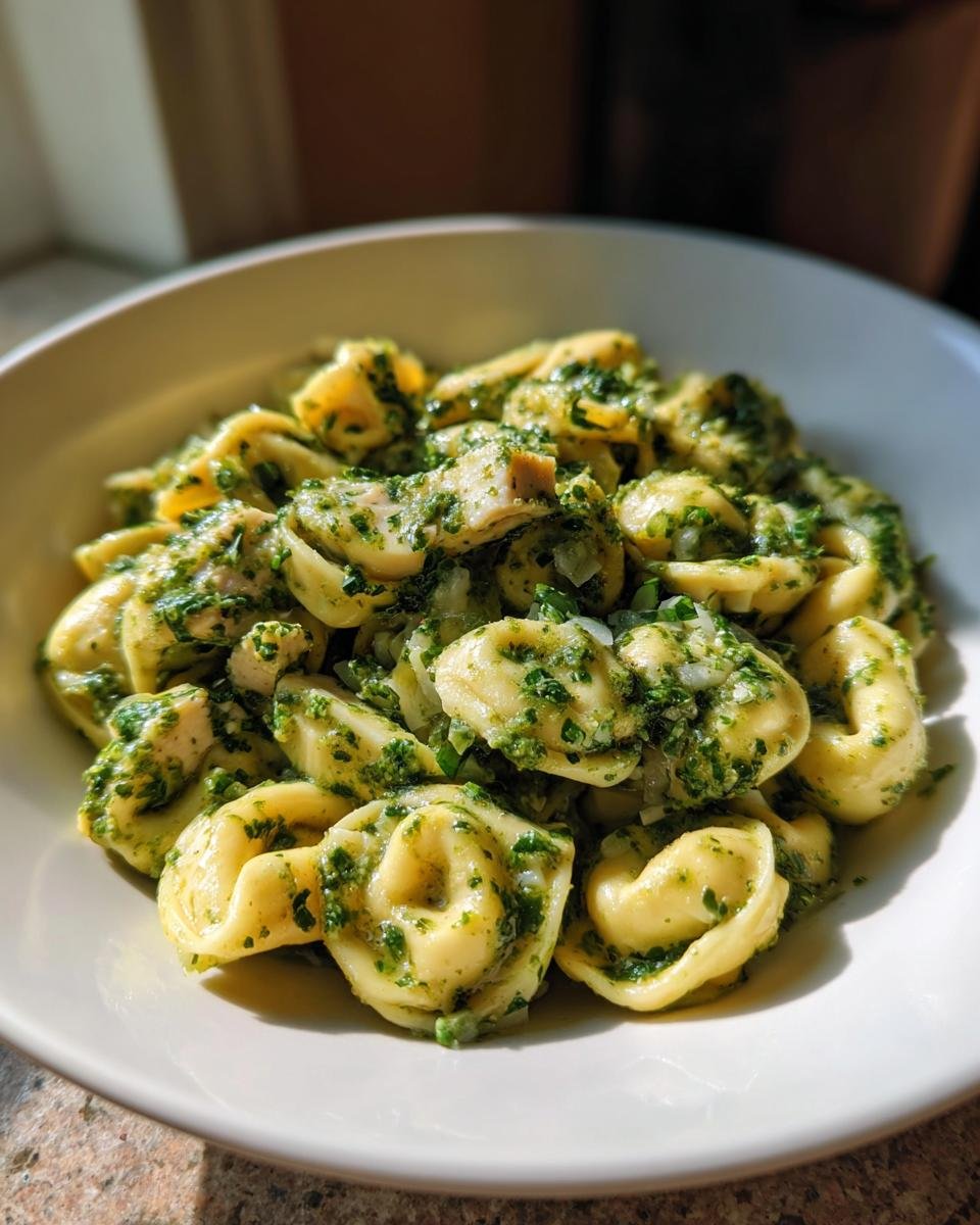 Close-up of a white bowl filled with Chicken Pesto Tortellini coated in a vibrant green pesto sauce.