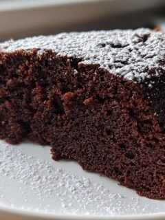 A close-up of a rich, dark slice of Chocolate Olive Oil Cake dusted generously with powdered sugar on a white plate.