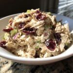 A close-up shot of creamy Cranberry Chicken Salad featuring shredded chicken, dried cranberries, celery, and red onion in a white bowl.