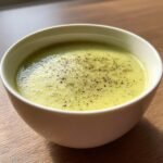 A close-up of a white bowl filled with smooth, light green Cream Zucchini Soup, topped generously with freshly ground black pepper.
