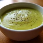 Close-up of a white bowl filled with smooth, light green Cream Zucchini Soup, topped with cracked black pepper.