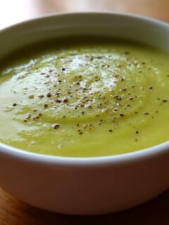 Close-up of a white bowl filled with smooth, light green Cream Zucchini Soup, topped with cracked black pepper.