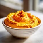 A close-up of bright orange Creamy Carrot Hummus swirled in a white bowl, drizzled with olive oil and sprinkled with paprika.