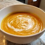 Close-up of a white bowl filled with vibrant orange Creamy Carrot Soup, garnished with a swirl of white cream.