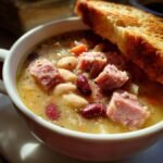 Close-up of a bowl of creamy Ham And Bean Soup featuring chunks of ham, white beans, and kidney beans, served with toasted bread.