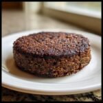 A close-up of a thick, dark brown, perfectly seared Easy Black Bean Burger patty resting on a white plate.