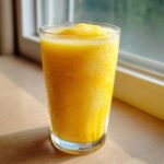 A tall glass filled with bright yellow, icy Frosted Lemonade sitting on a wooden surface near a window.
